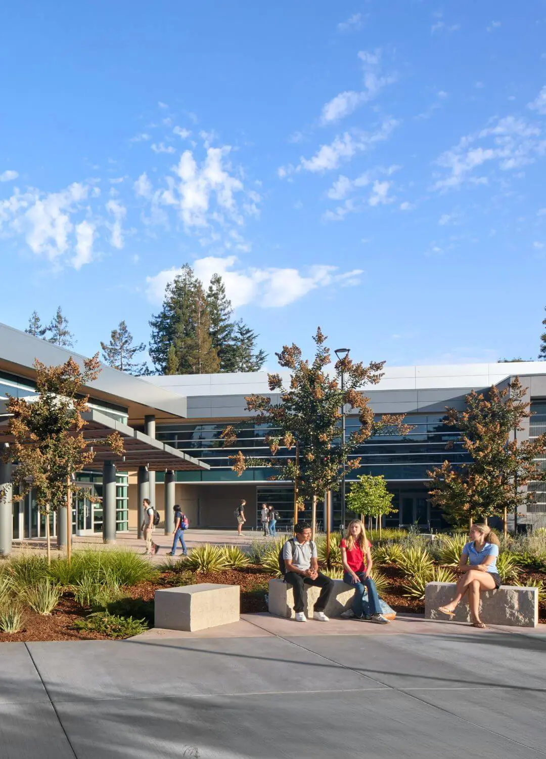 A group of friends gathered in the Quad outside of the Welcome Center, sitting on benches and chatting happily in the sunshine, while other groups were walking through.