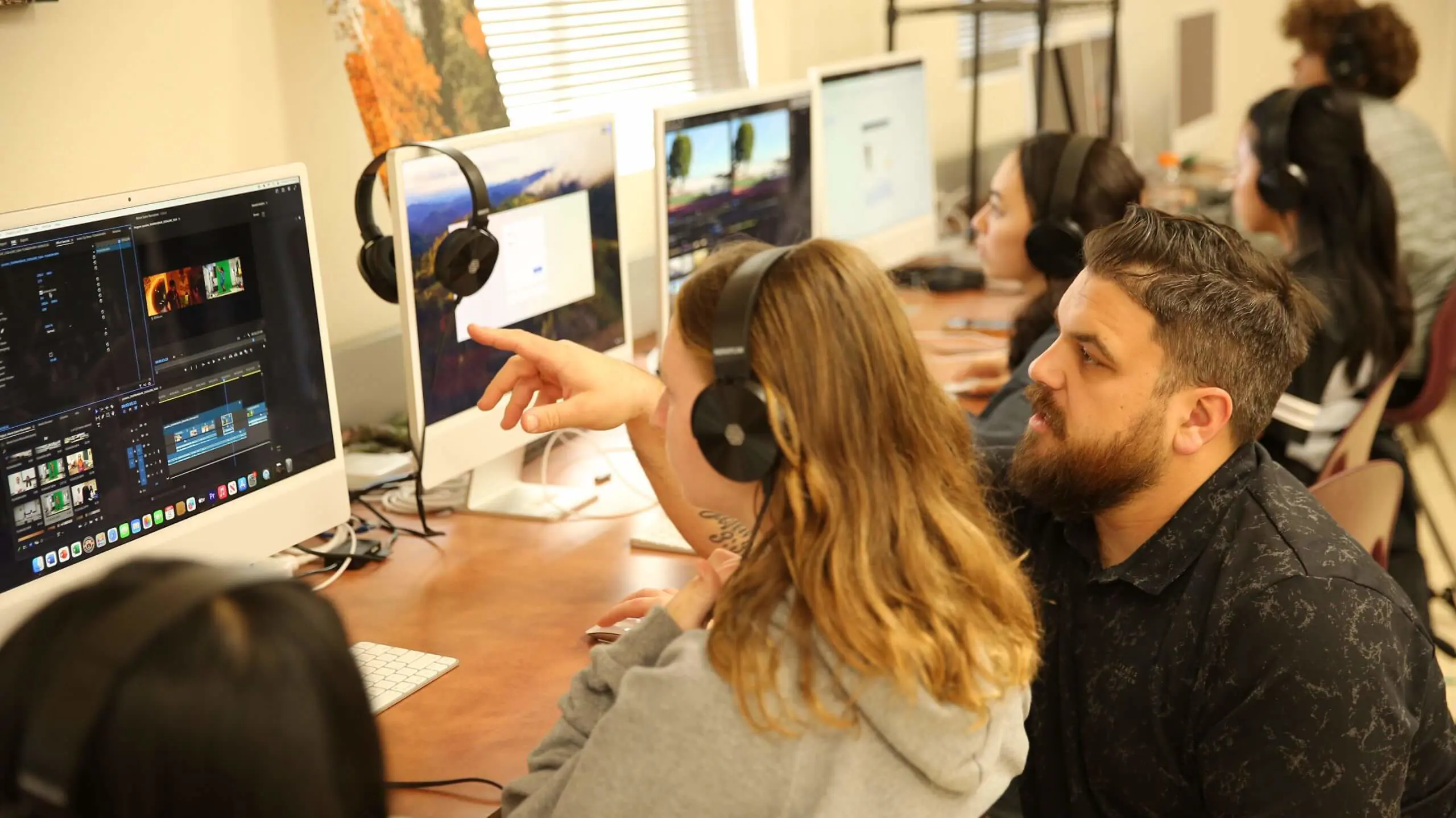 An educator guides a student in video editing on a desktop computer. Both wear headphones, focused. Other students work on similar projects in the background.