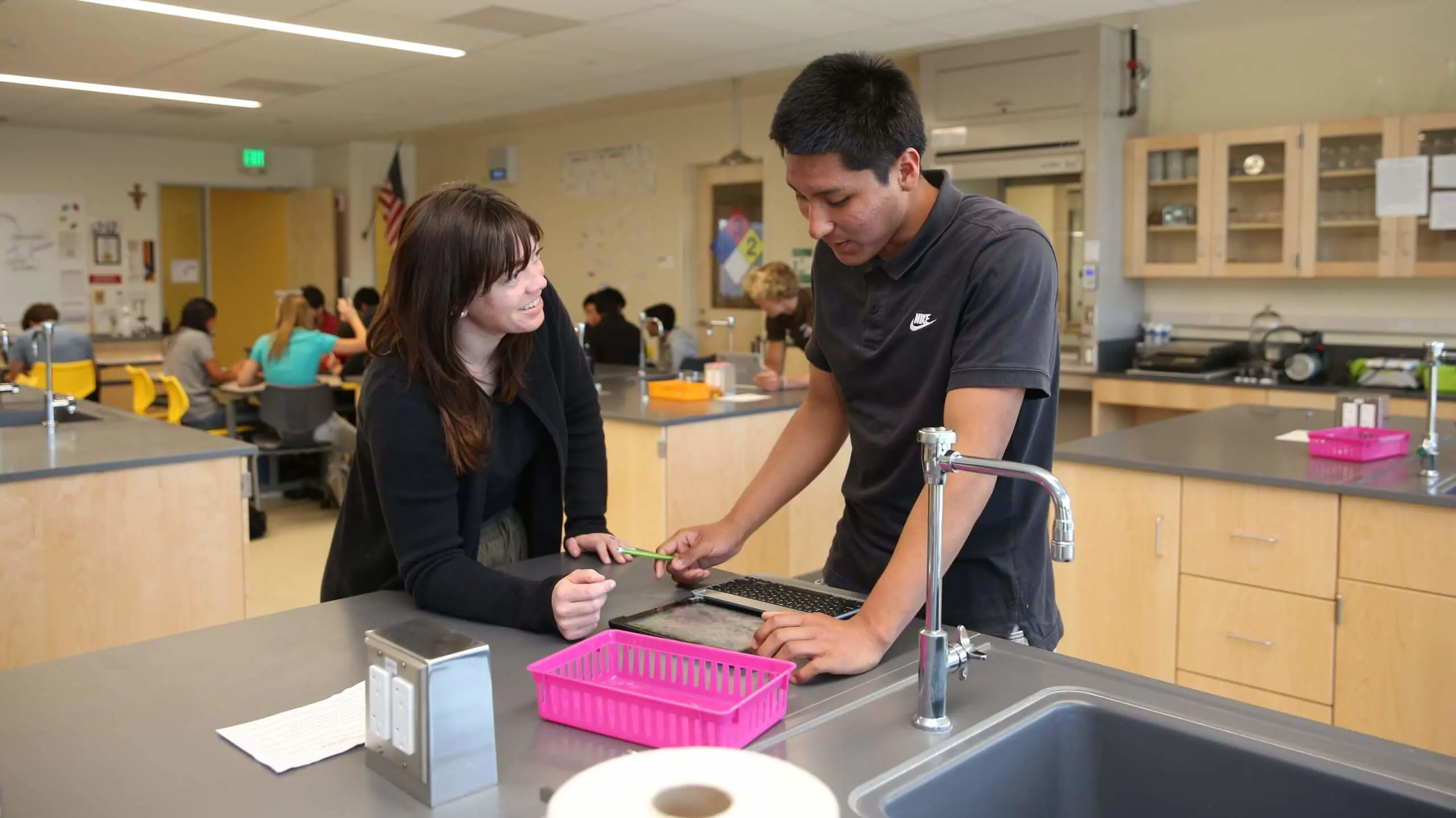 An educator and student engage in discussion over a tablet in a science classroom. Background shows other students at tables, with a colorful and collaborative atmosphere.