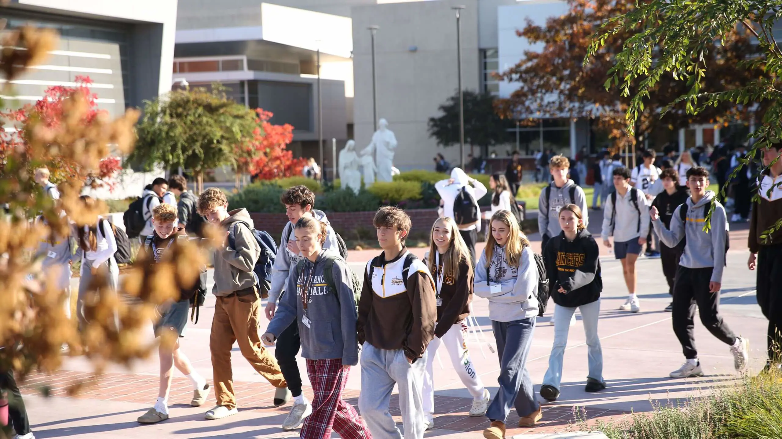 Lancer students walk across a sunlit campus with modern buildings and autumn trees in the background, creating a lively, energetic scene.