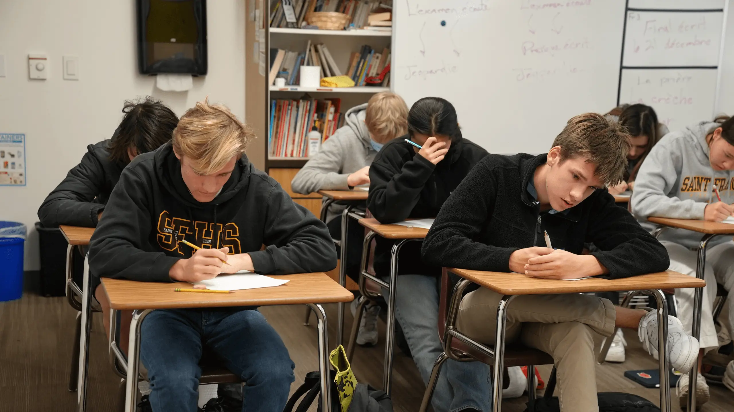 Students sit at wooden desks in a classroom, focused and writing on papers. Shelves of books and a whiteboard are visible in the background.