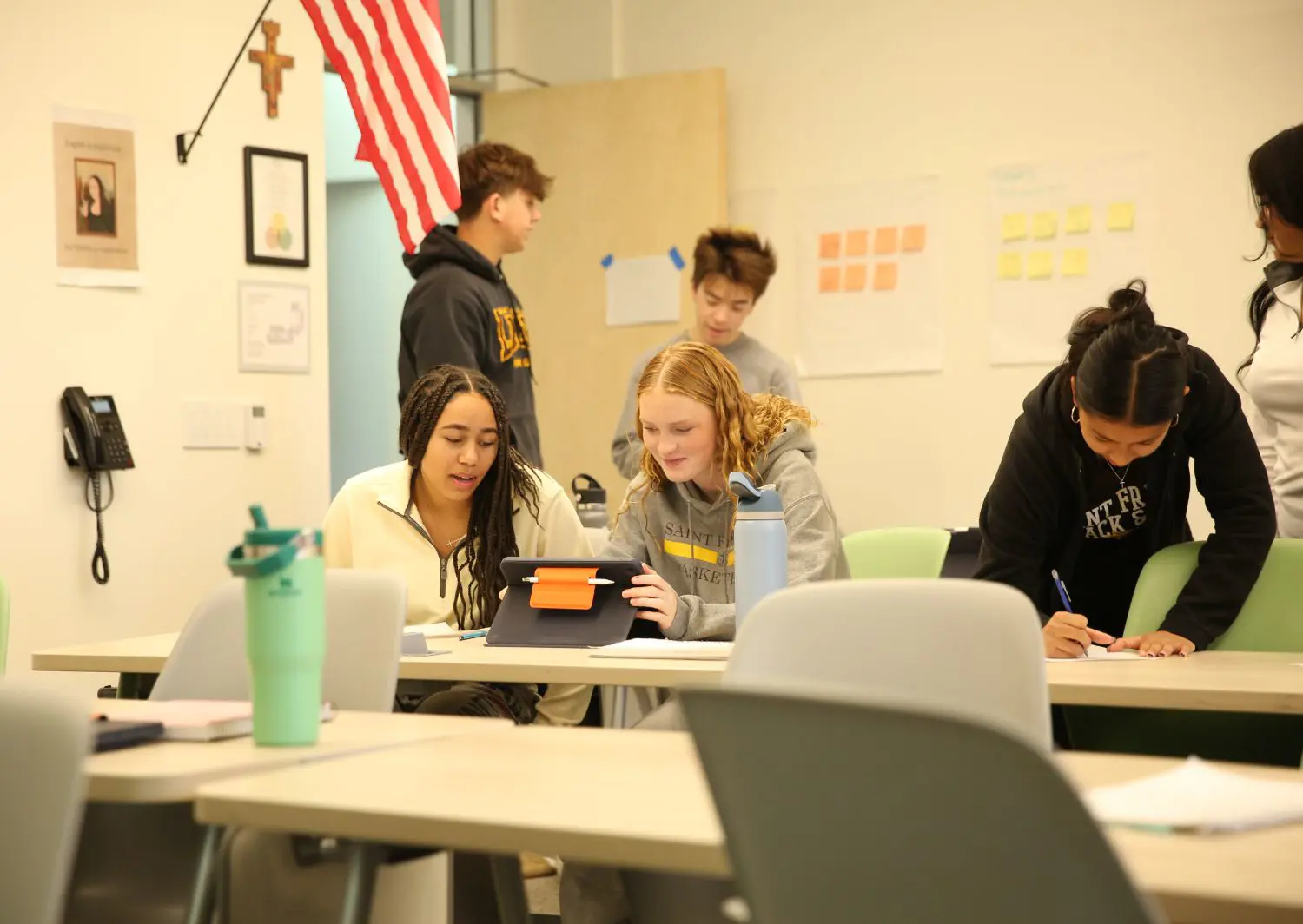 Students collaborate in a classroom, with two focused on a tablet, one taking notes. The setting is casual, with an American flag and posters visible.