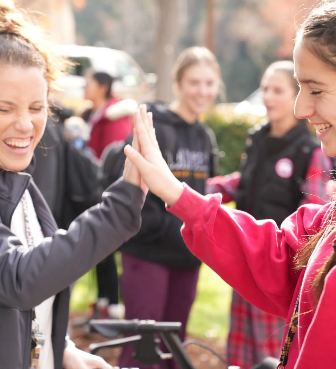 An educator and student sharing a high-five outdoors, surrounded by a blurred group in casual attire. The mood is cheerful and energetic.