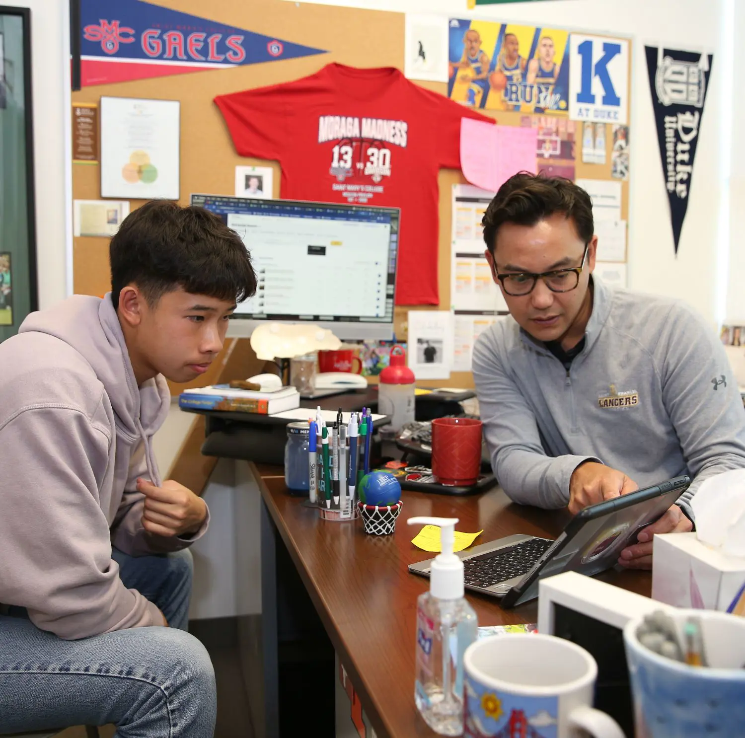 A student and counselor sit in an office filled with sports memorabilia and college banners. They are focused on a tablet together, creating an engaged and collaborative atmosphere.
