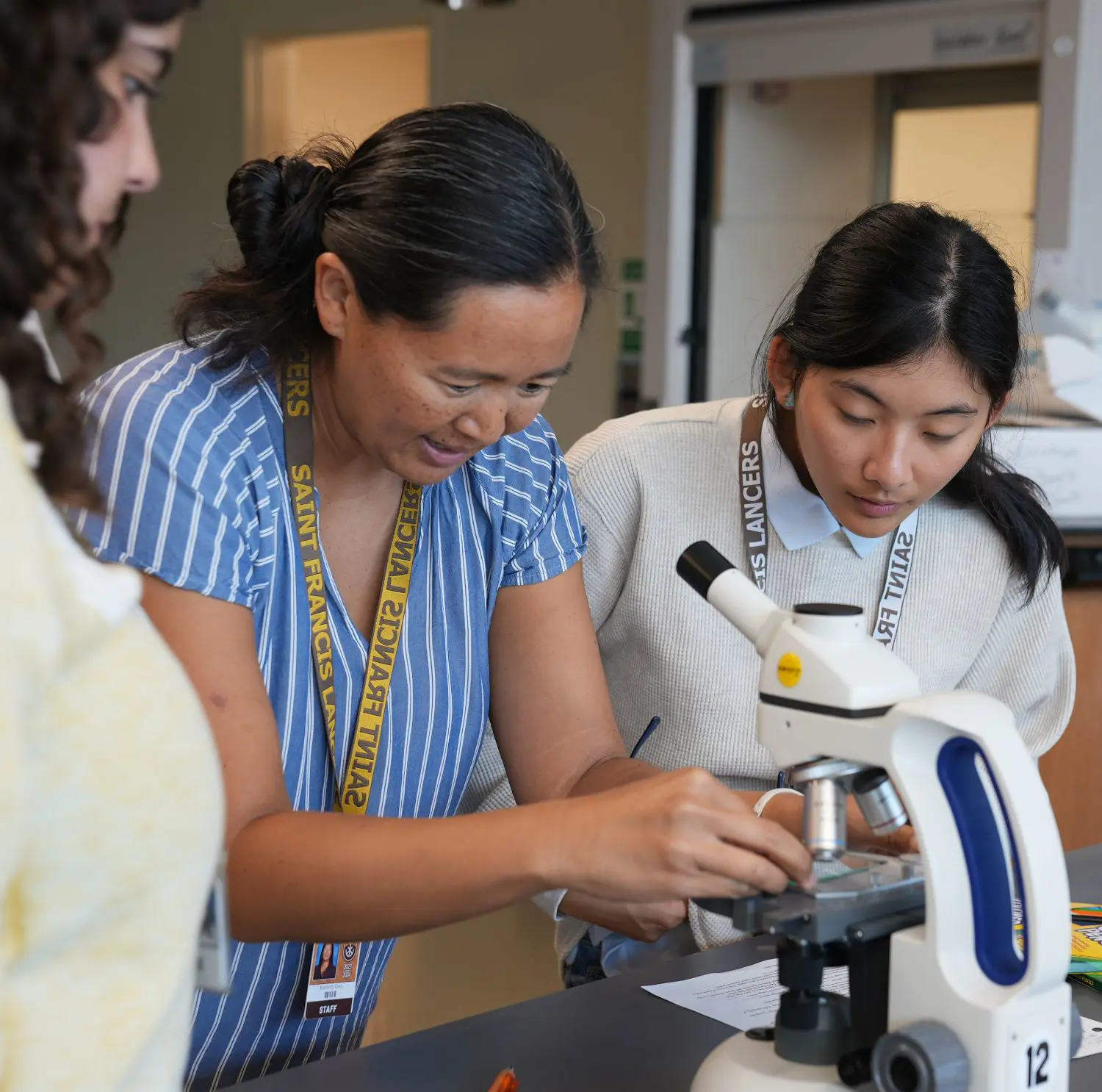 An educator and a student focus intently on adjusting a microscope in a bright classroom. Nearby are lab materials like a beaker and markers.