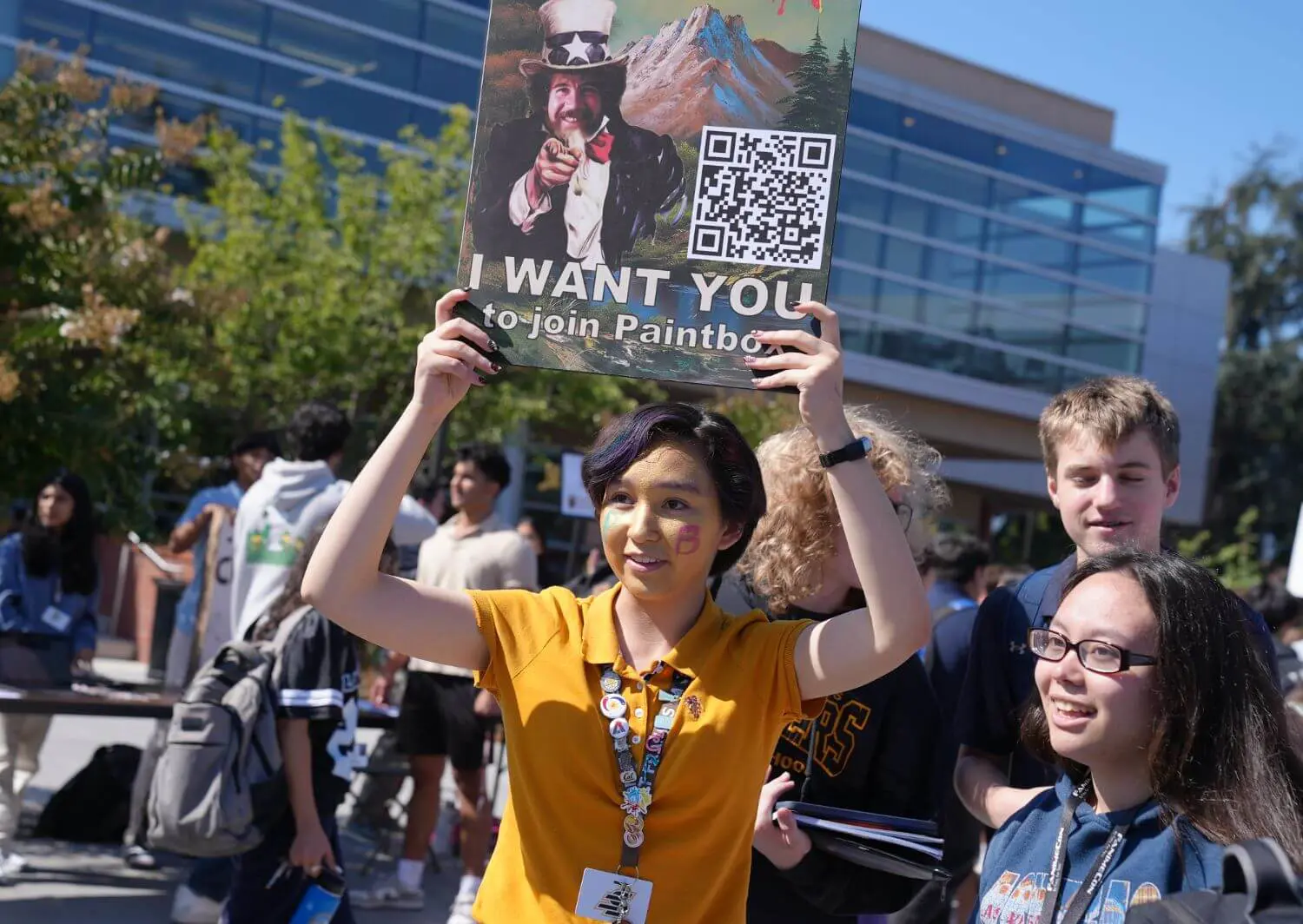 Student at the annual activity fair holding up a sign saying “I want you” with a depiction of Bob Ross as Uncle Sam to get people to sign up for Paintbox club.