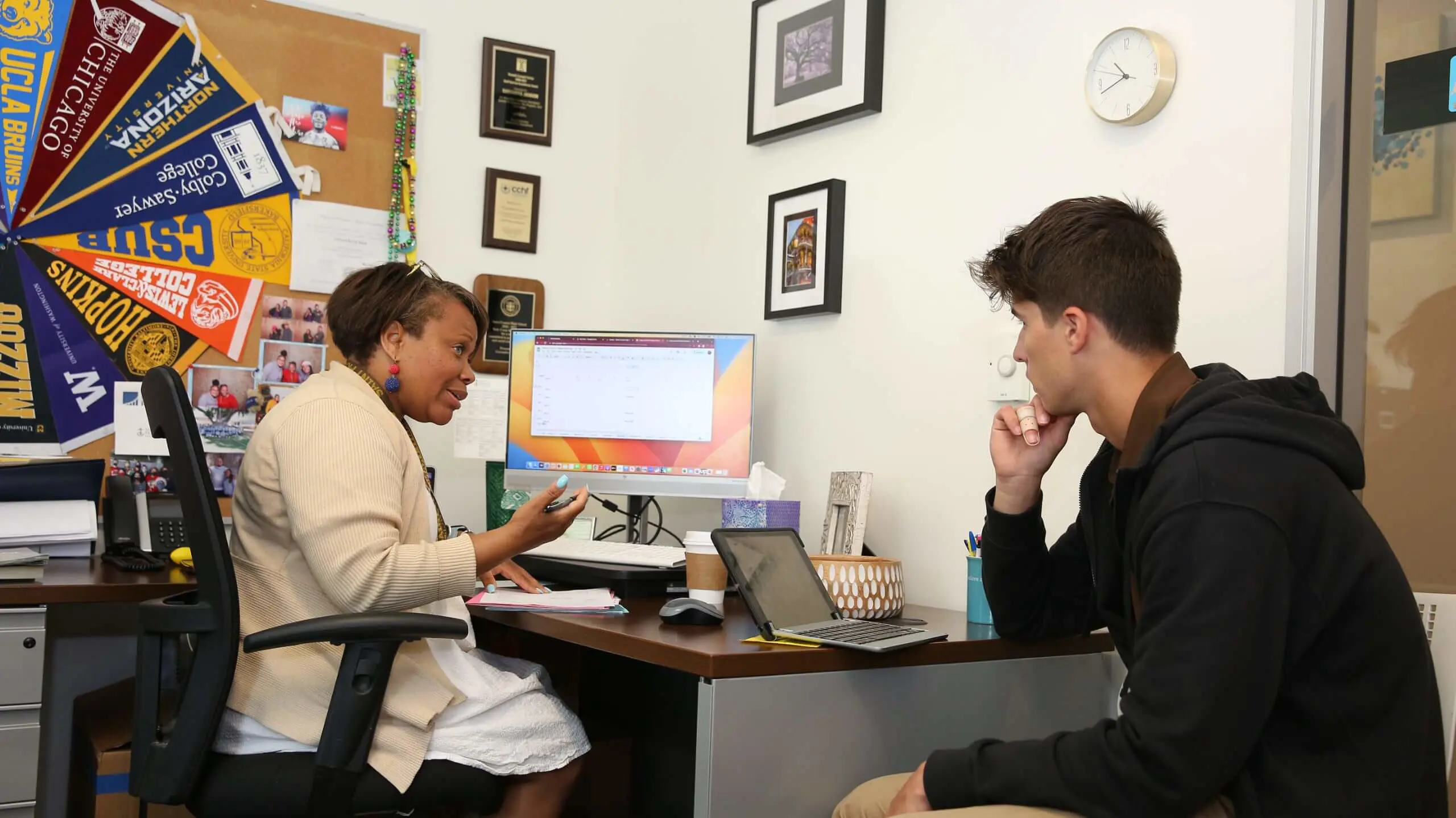 A counselor and a young man sit across a desk in an office. The counselor gestures while talking, and the man listens intently. College pennants adorn the wall.