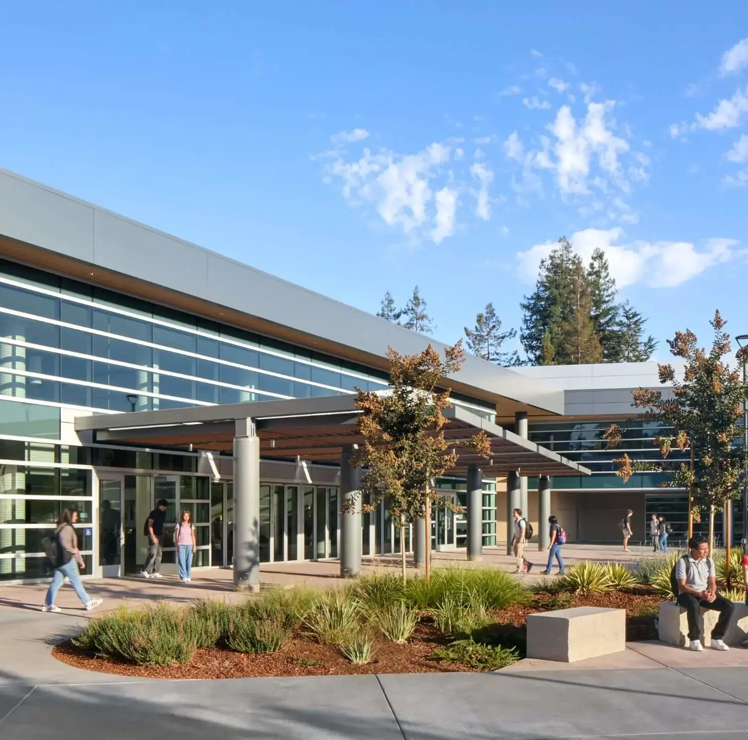 A group of friends gathered in the Quad outside of the Welcome Center, sitting on benches and chatting happily in the sunshine, while other groups were walking through.
