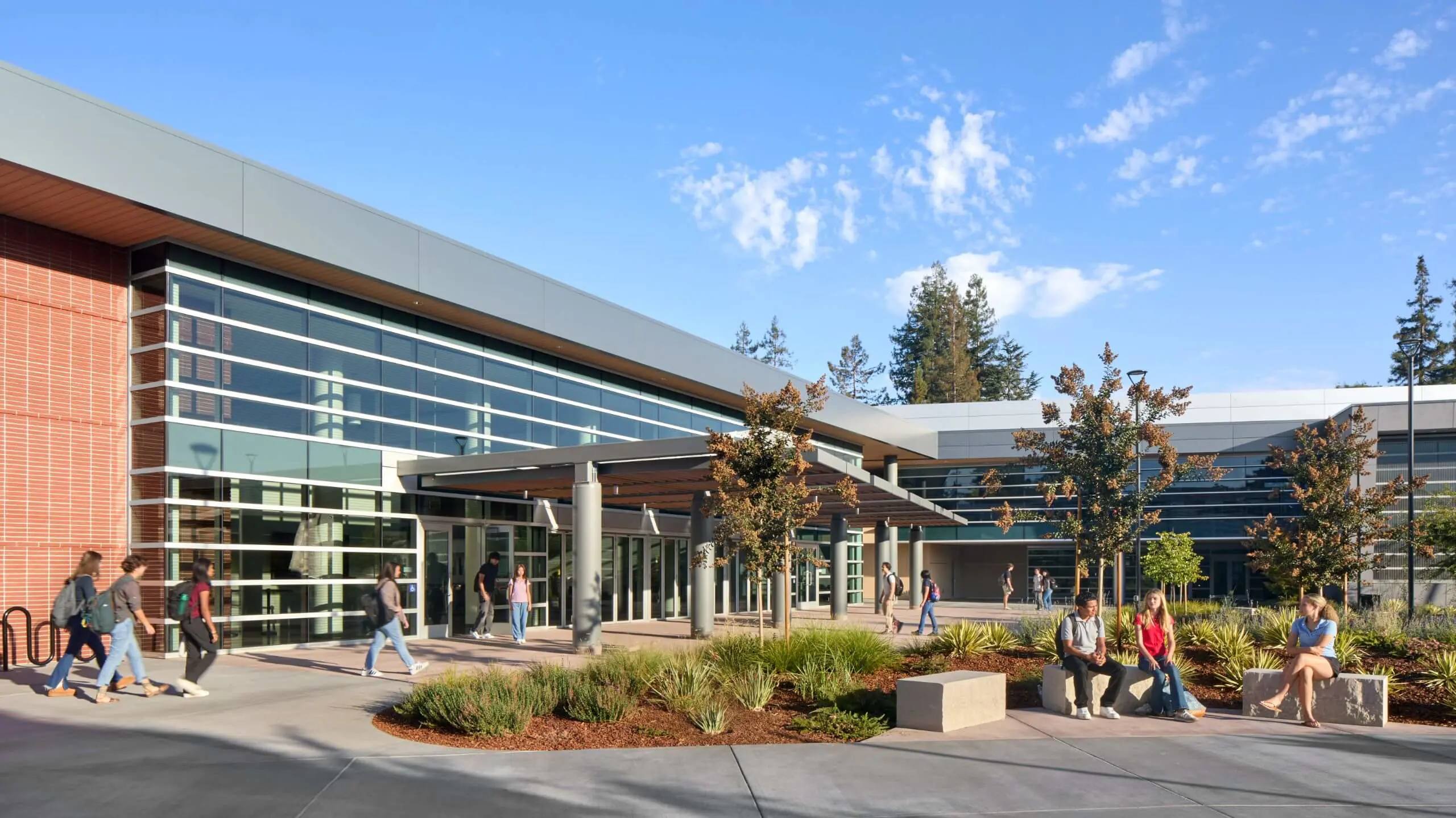 A group of friends gathered in the Quad outside of the Welcome Center, sitting on benches and chatting happily in the sunshine, while other groups were walking through.