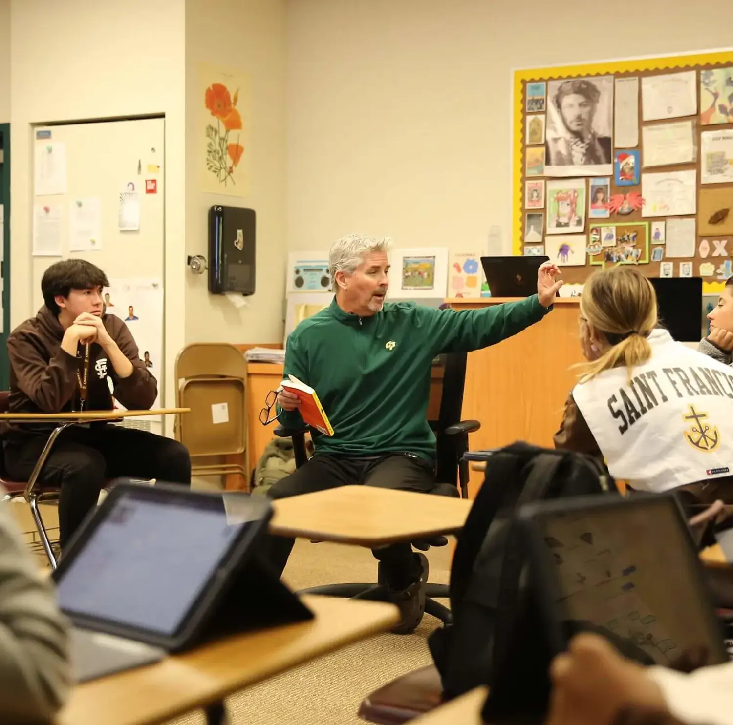 An educator in a green shirt engages students in a classroom. Some students use tablets, and the walls are decorated with various educational materials and artwork.