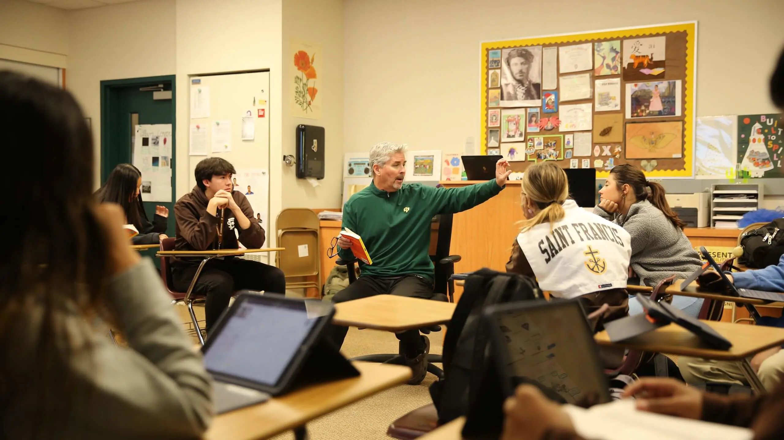 An educator in a green sweater passionately gestures while holding a book, engaging students in a classroom. Students sit attentively with tablets and notebooks.