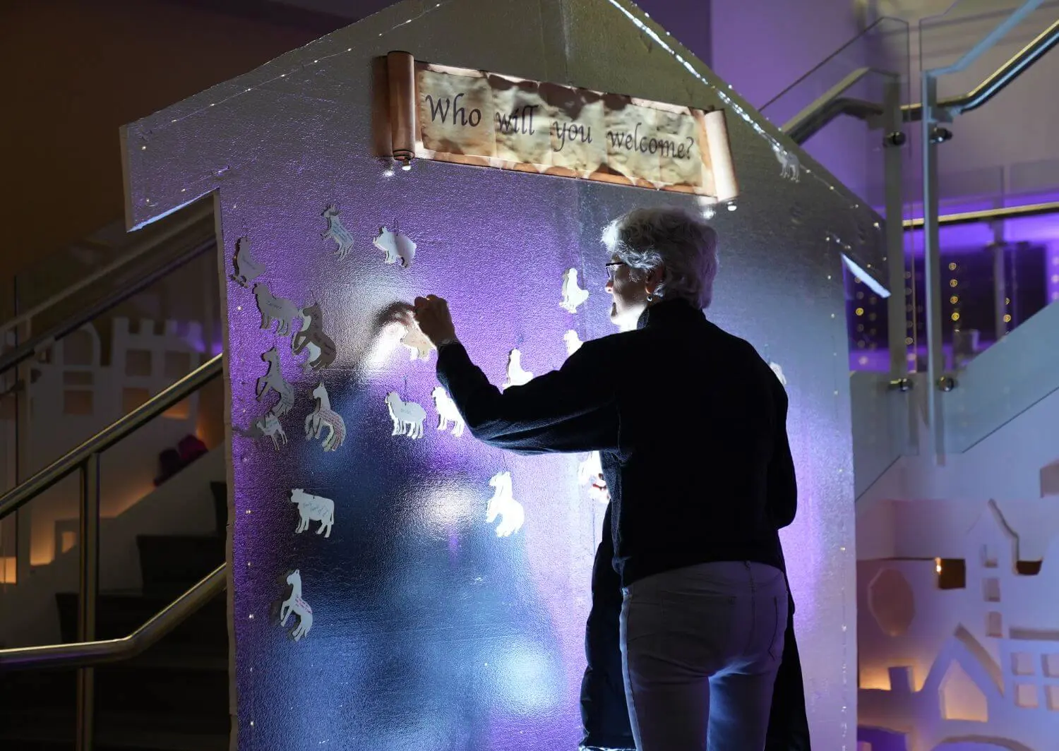 A woman interacts with an illuminated display featuring animal cutouts and a sign that reads "Who will you welcome?" during an Advent experience in a beautifully decorated indoor space.