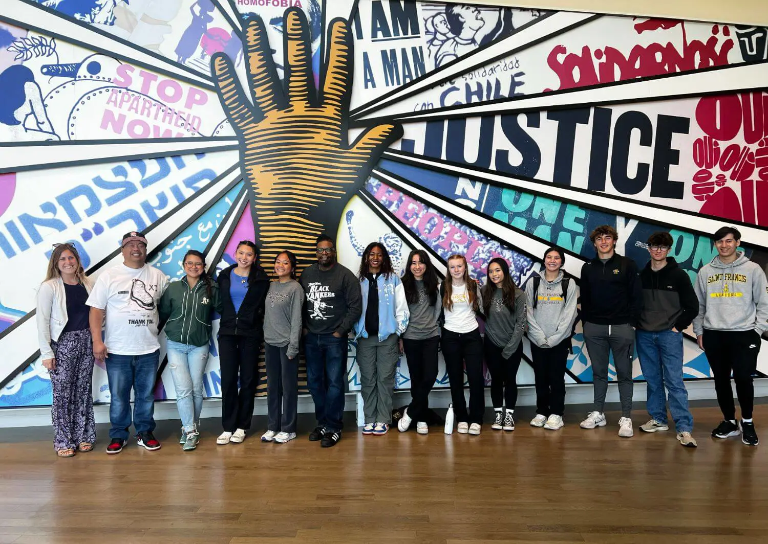 A group of students and educators pose in front of a vibrant mural featuring a large hand and words promoting justice, solidarity, and activism.