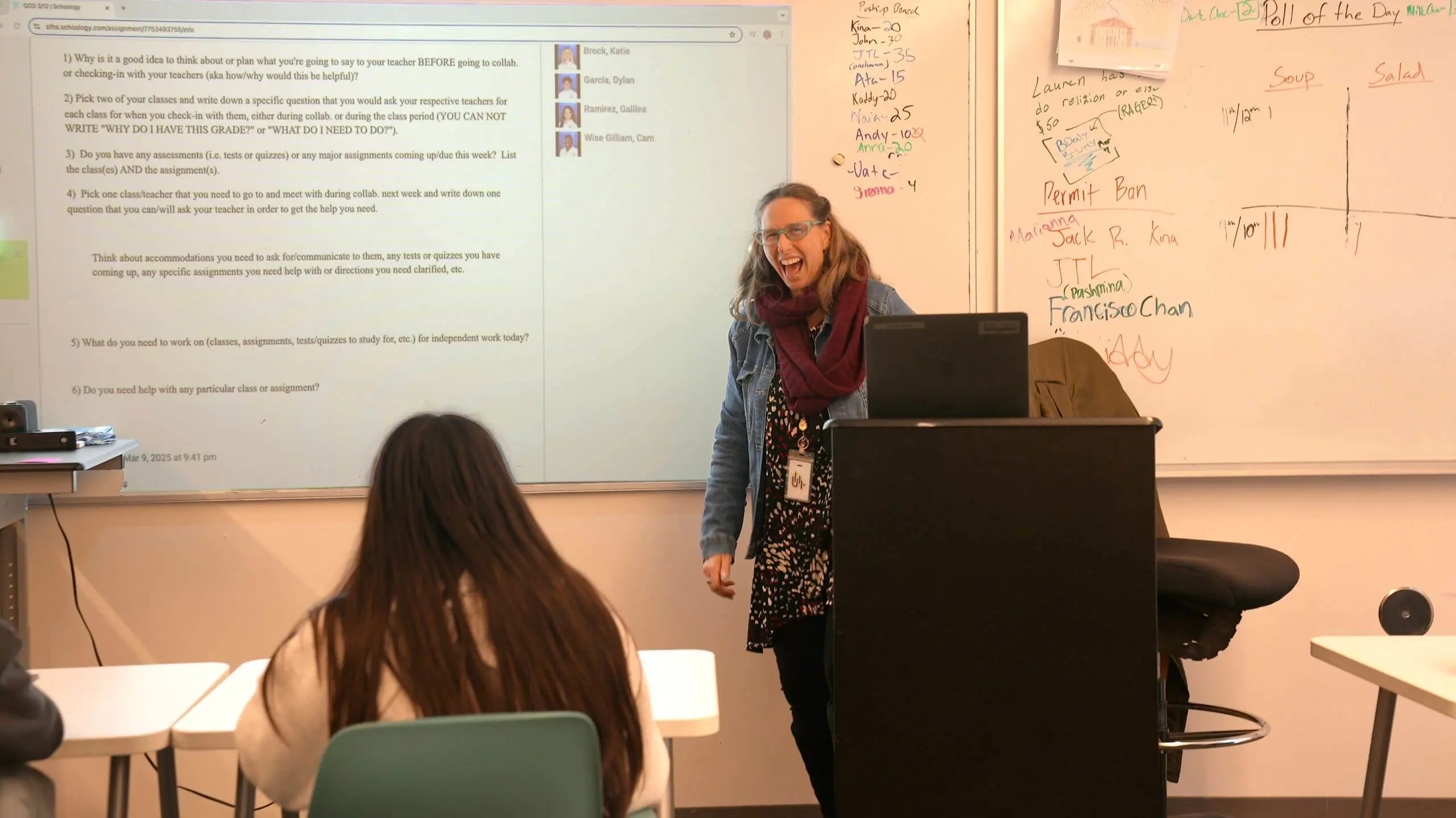 An educator stands at the front of a classroom, engaging students. The projector displays class instructions, and a whiteboard lists names and a poll.