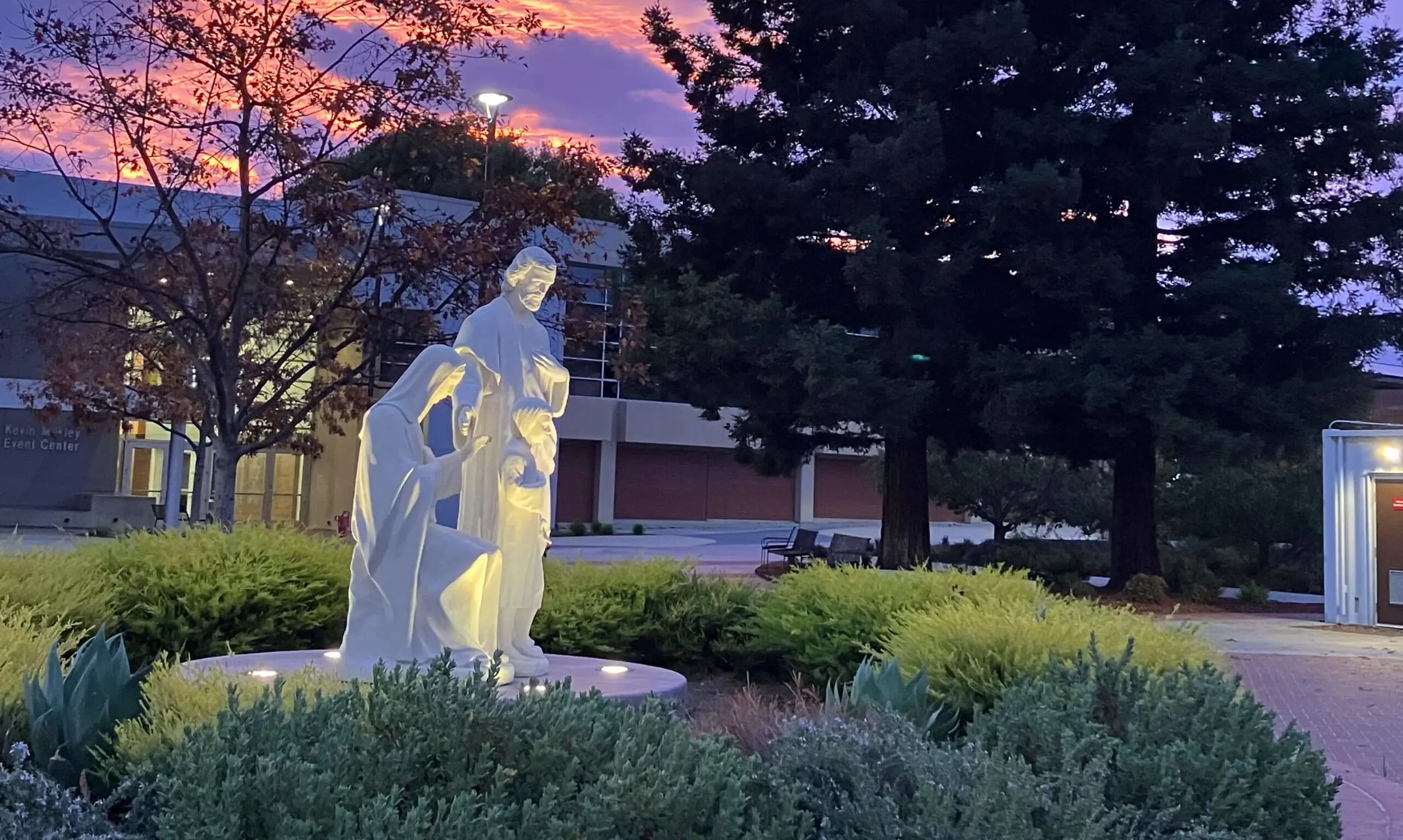 A white statue of the Holy Family is illuminated in a landscaped garden, set against a stunning sunrise with vibrant pink, orange, and purple hues in the sky.