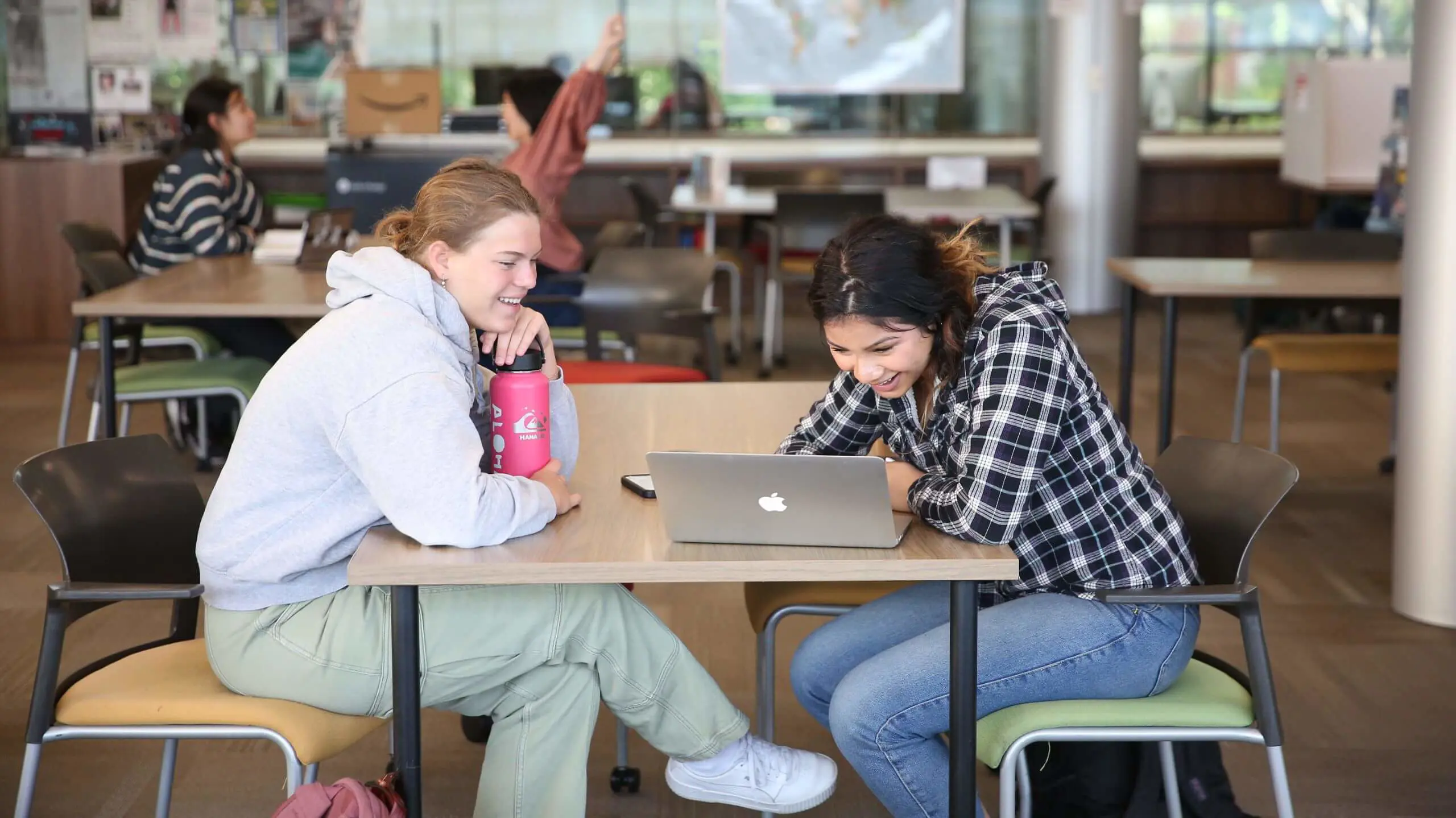 Two smiling students sitting at a table looking at their laptops.