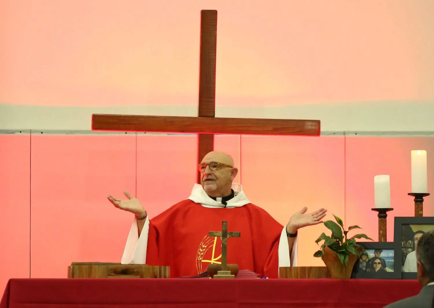 A priest wearing red vestments stands behind an altar with outstretched arms, leading a religious service in front of a wooden cross, candles, and a framed photo, with a congregation in attendance.