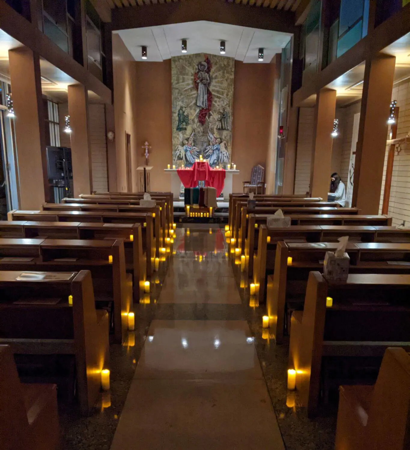 A dimly lit chapel at night features an illuminated aisle lined with glowing candles, leading to an altar adorned with red cloth and candles, with a single person praying in the pews.