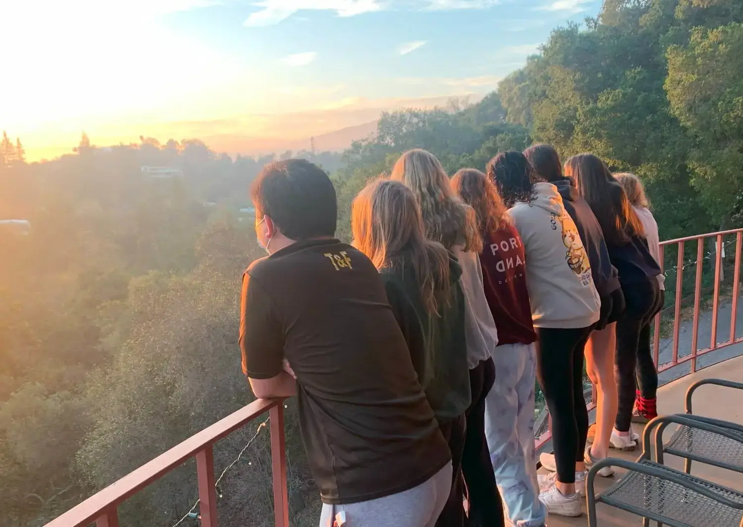 A group of students on Senior Retreat stand together on a balcony, leaning against the railing as they watch a sunrise over a lush, tree-covered landscape.