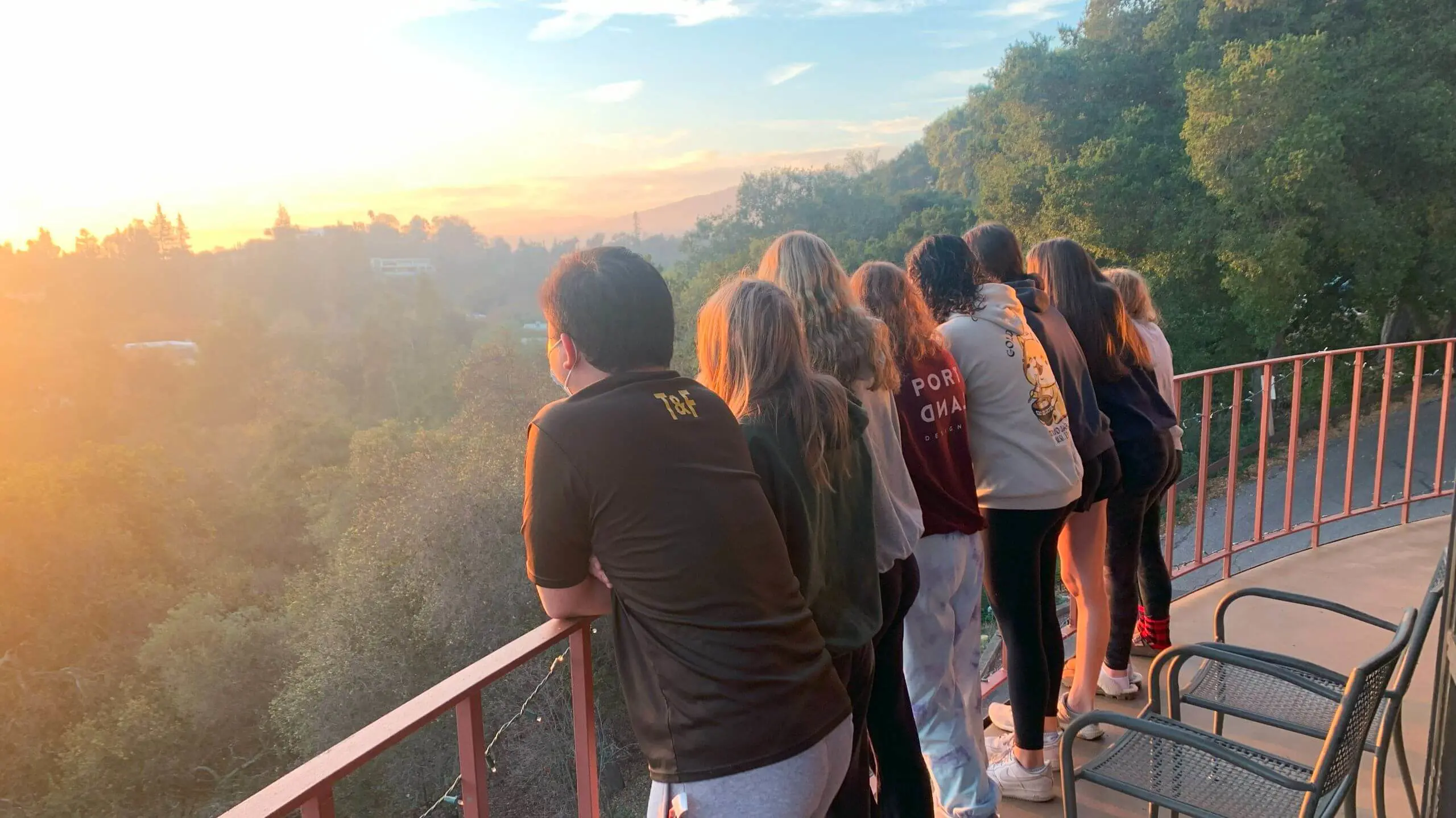 A group of students on Senior Retreat stand together on a balcony, leaning against the railing as they watch a sunrise over a lush, tree-covered landscape.