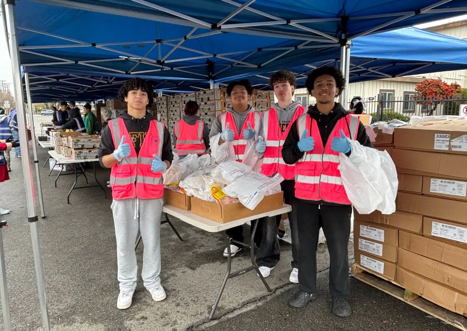 A group of young student volunteers wearing pink safety vests and blue gloves, stand under blue canopy tents at an outdoor food distribution event, giving thumbs-up while handling bags of supplies.