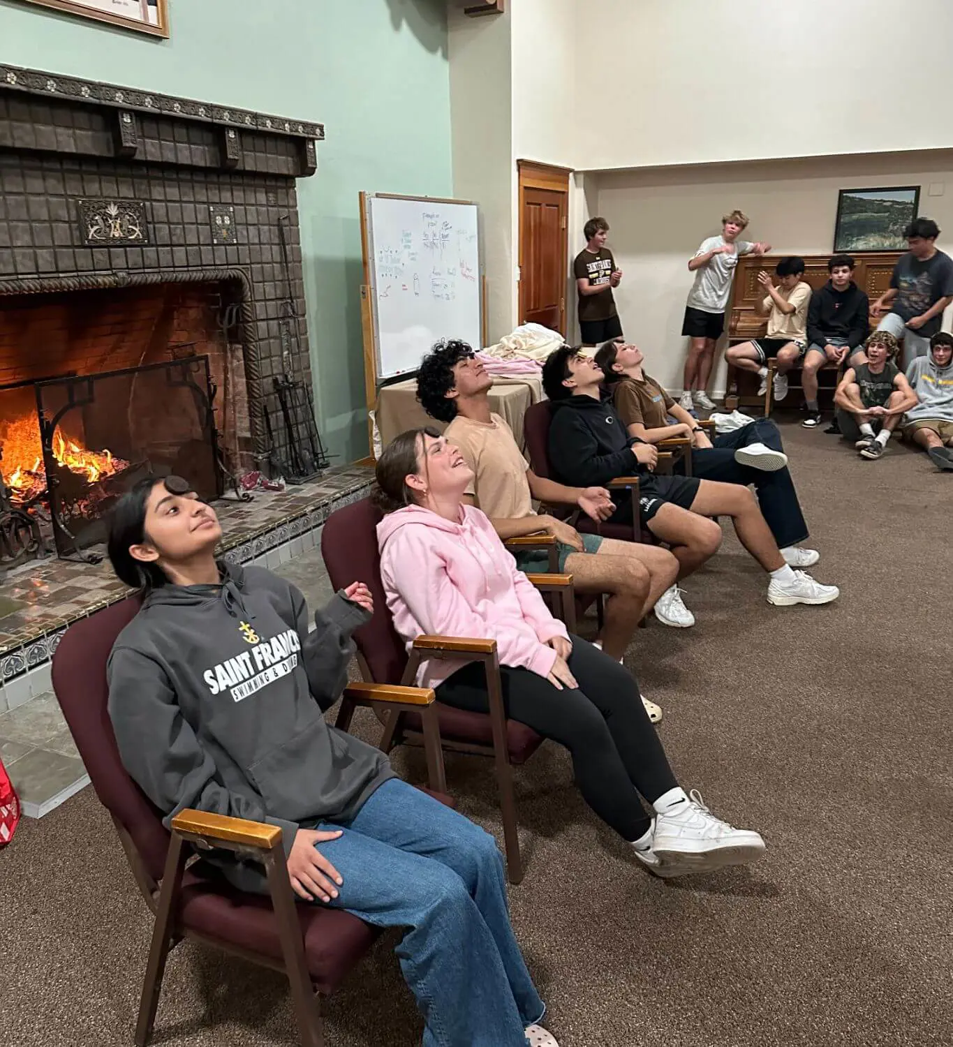 The photo shows a group of students in a cozy lodge-like setting, with some seated in chairs participating in an Oreo cookie challenge while others watch and laugh near a fireplace.