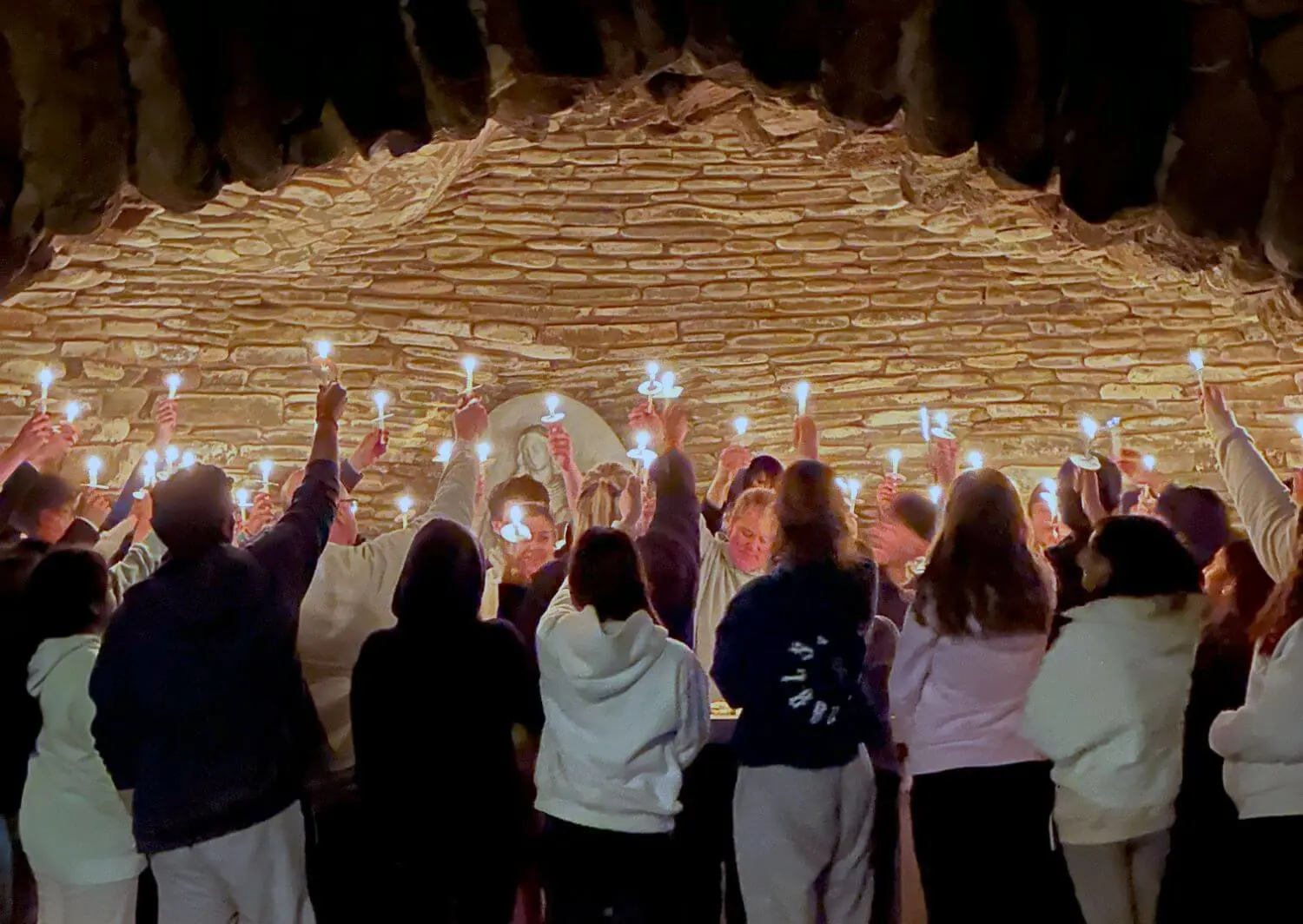 A group of senior students stand together in a grotto at night, holding up lit candles in a solemn and reflective gathering.