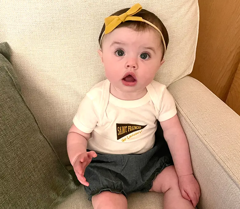 A baby girl wearing a shirt featuring a Little Lancers Flag, smiling and sitting on a soft surface