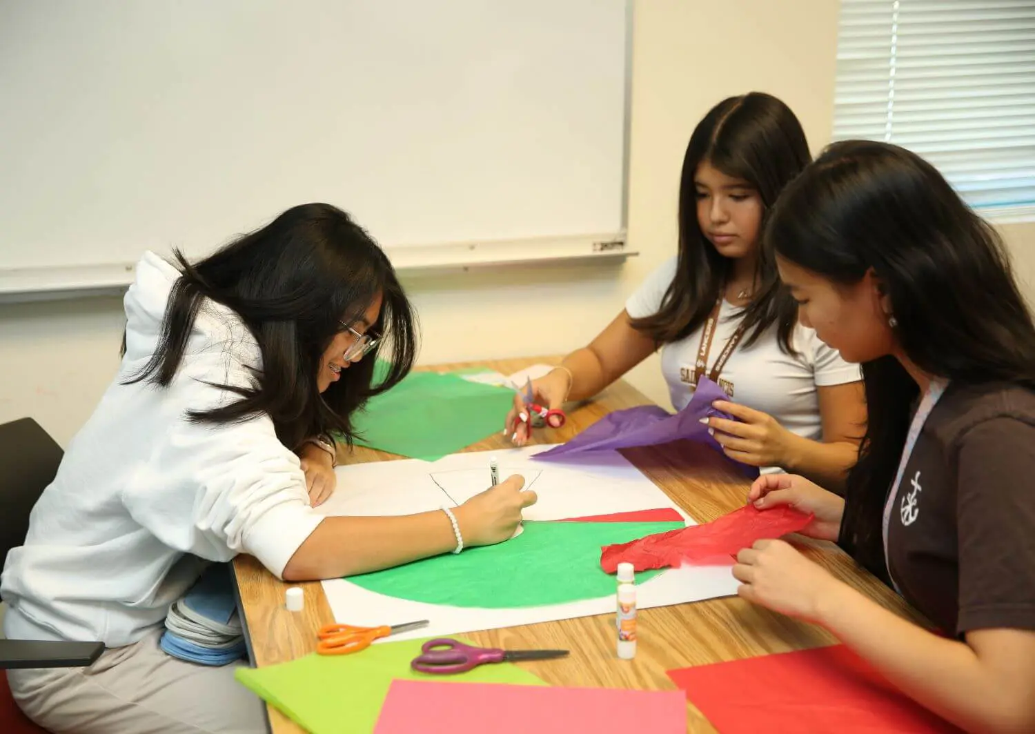 Three students engaged in an art project at a wooden table, using colored paper, glue, and scissors. The scene conveys creativity and concentration.