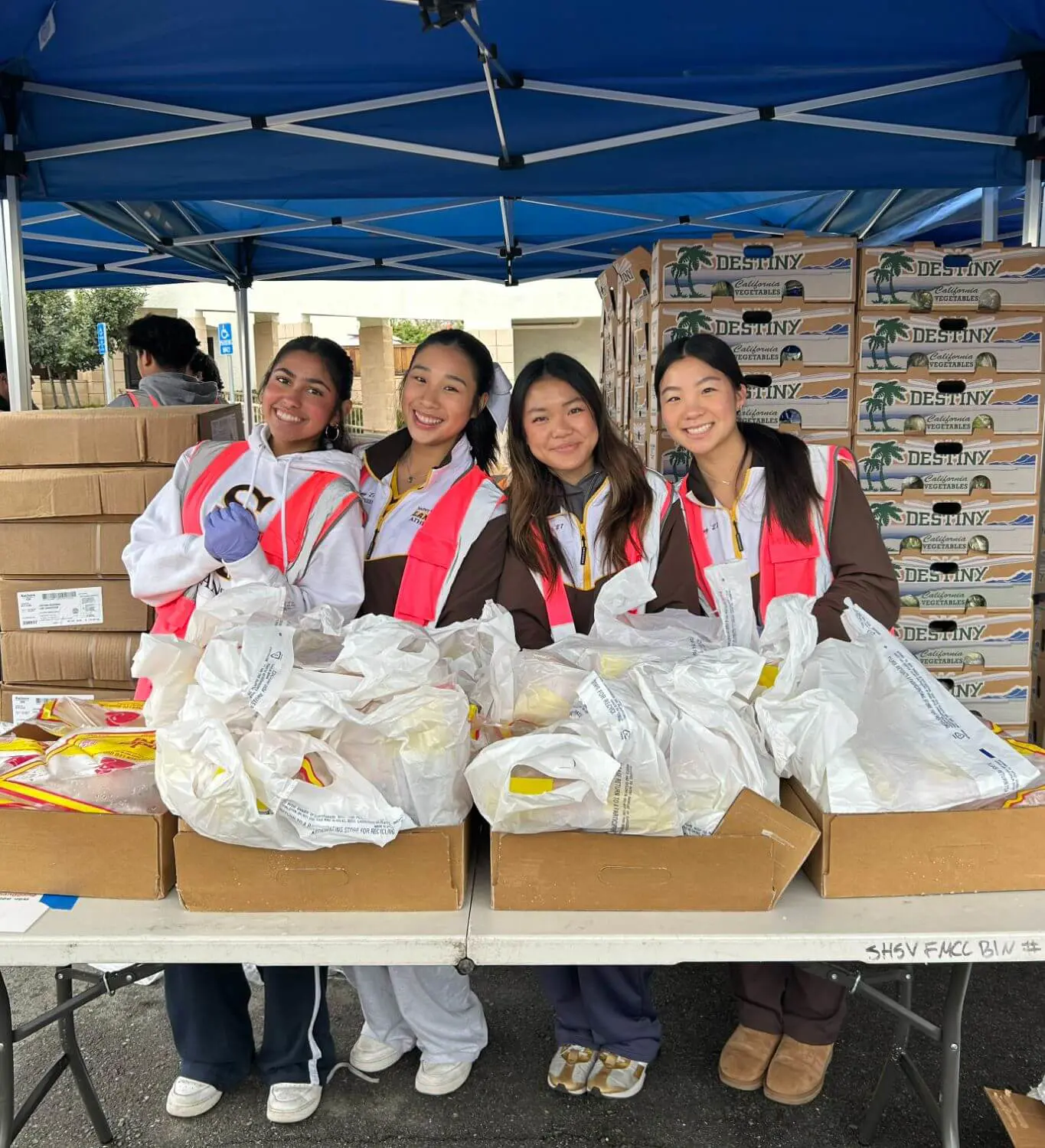 Four young Lancer women wearing safety vests are smiling while volunteering at a food distribution event under a tent, surrounded by boxes of supplies and bags of food.