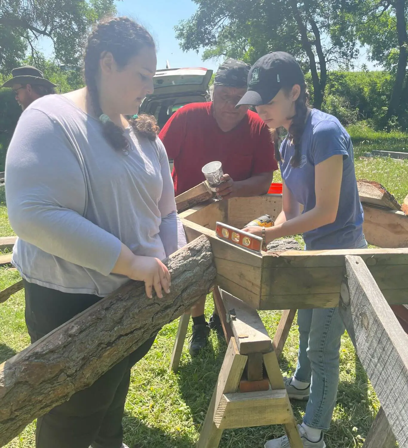 A group of students work together outdoors on a wooden construction project, using tools and a level while handling logs and wooden frames.