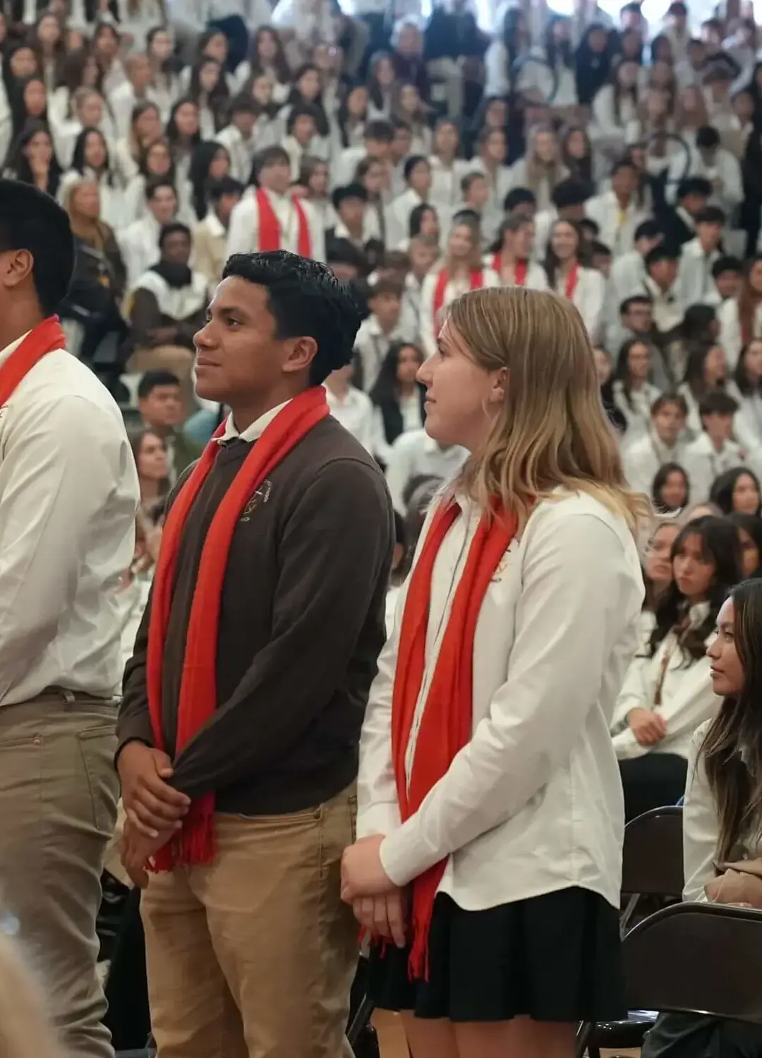 Students dressed in their white liturgy shirts, with three standing in the foreground wearing red scarves, during an introduction of confirmandi in Burns gymnasium. 