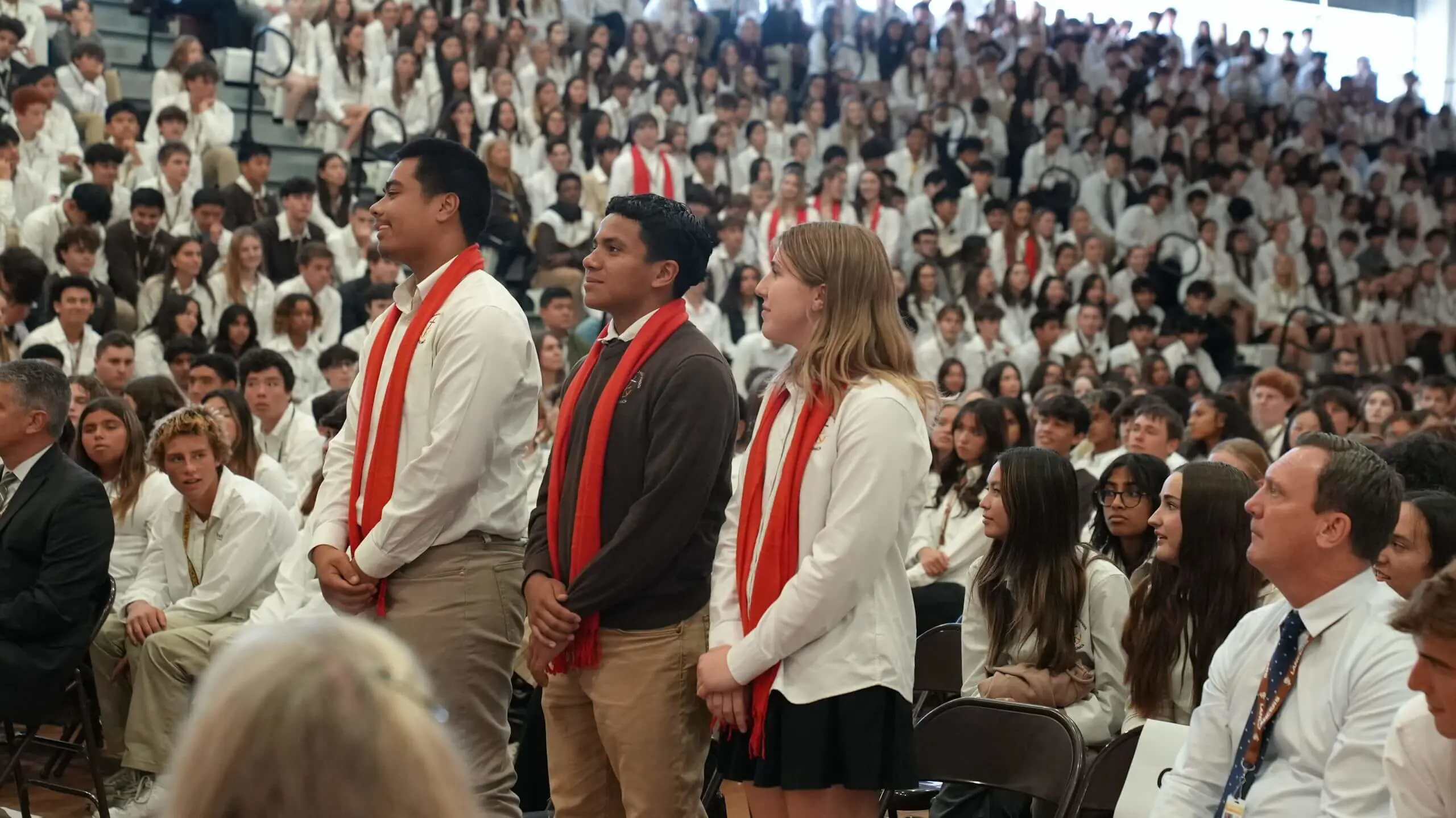 Students dressed in their white liturgy shirts, with three standing in the foreground wearing red scarves, during an introduction of confirmandi in Burns gymnasium.