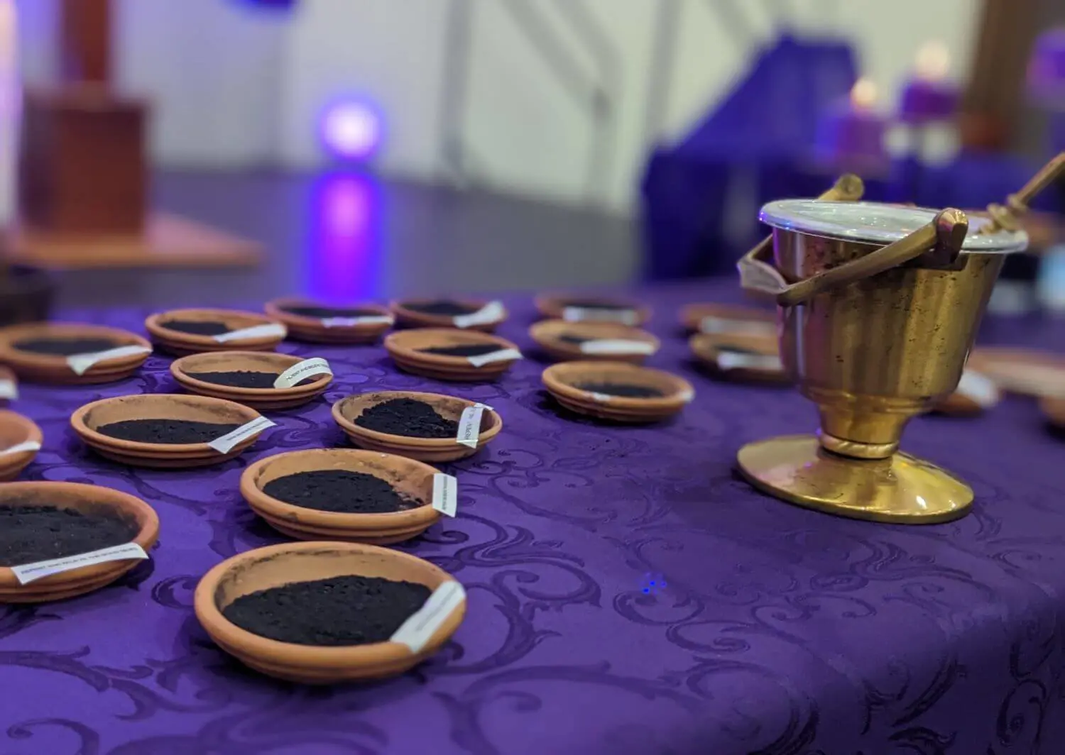 The photo shows small clay dishes filled with ashes on a purple cloth-covered table, alongside a golden holy water bucket and sprinkler, prepared for the Ash Wednesday prayer service.