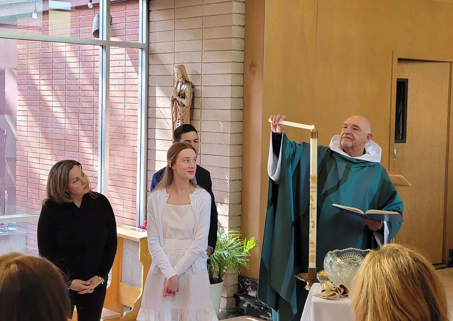 The photo captures a religious ceremony in the Chapel of Saint Joseph where Father Tony Mancuso is lighting a tall candle in front of a young woman in a white dress.