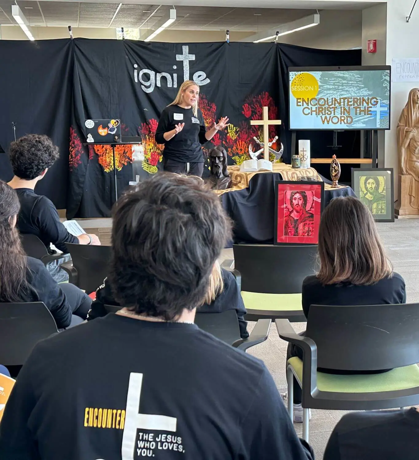 A speaker addresses a group of students at the Encounter Retreat, with a backdrop reading "Ignite" and a theme of "Encountering Christ in the Word," surrounded by religious imagery and artifacts.
